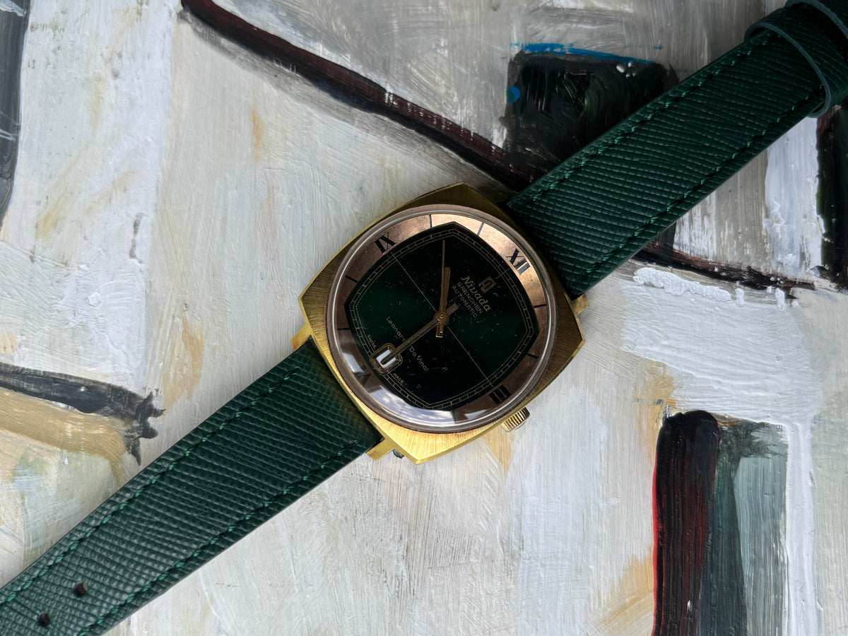 A vintage-style wristwatch with a gold-toned case, dark green dial, and a dark green woven strap, laid diagonally on a textured, painted off-white background.