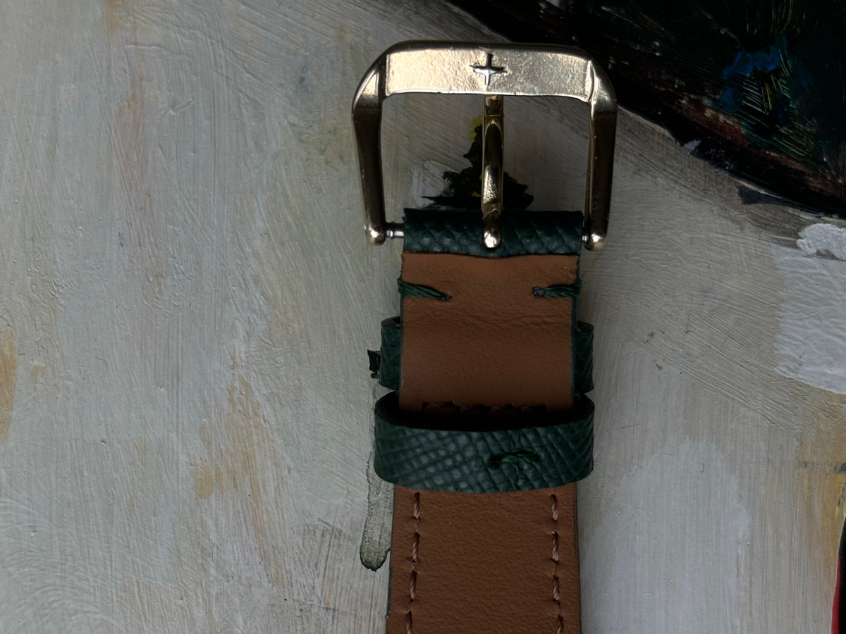 A close-up of a leather watch strap with a silver-toned metal buckle, featuring a tan center section and textured dark green outer layers, shown against a textured off-white background.