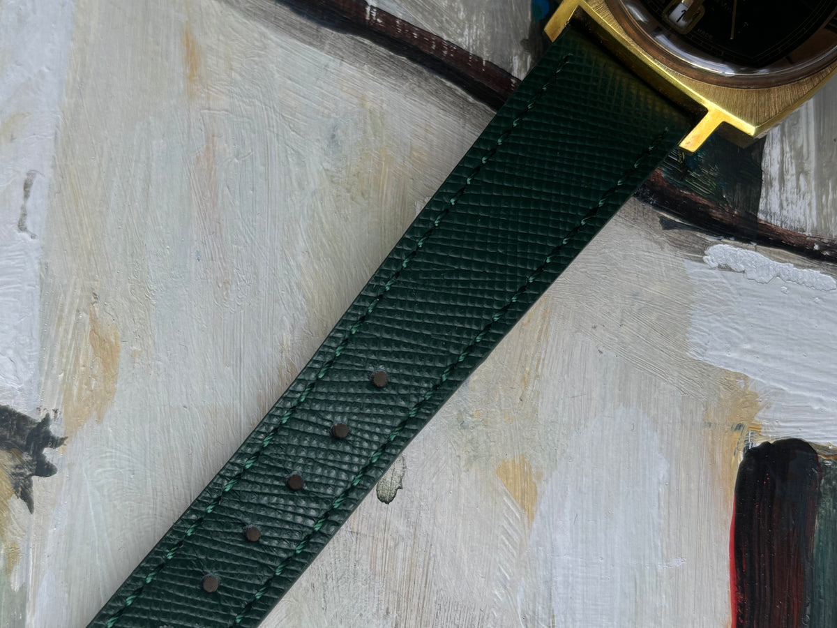 A close-up of a dark green textured leather watch strap attached to a gold-toned watch case, laid diagonally across a textured off-white painted surface.