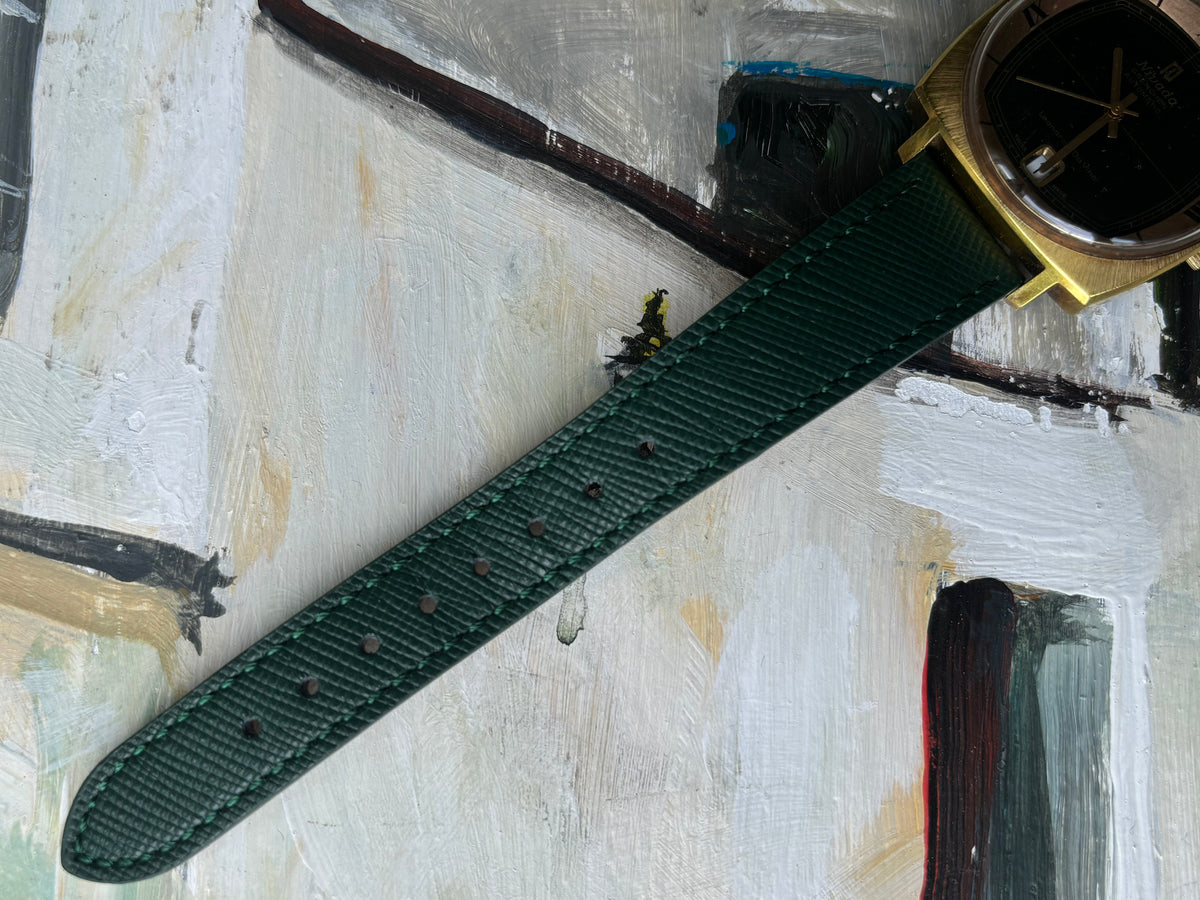 A close-up of a dark green textured watch strap attached to a gold-tone watch case, laid diagonally across an abstract painted background.