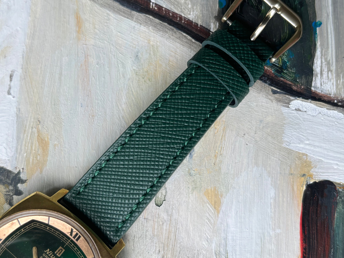 A close-up of a dark green textured leather watch strap with gold-toned hardware, laid diagonally across an abstract painted background.