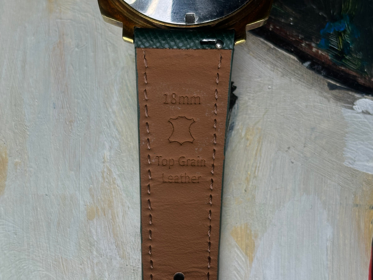 Close-up of a brown leather watch strap with visible stitching and embossed text reading '18mm' and 'Top Grain Leather', attached to a watch case against a textured off-white background.