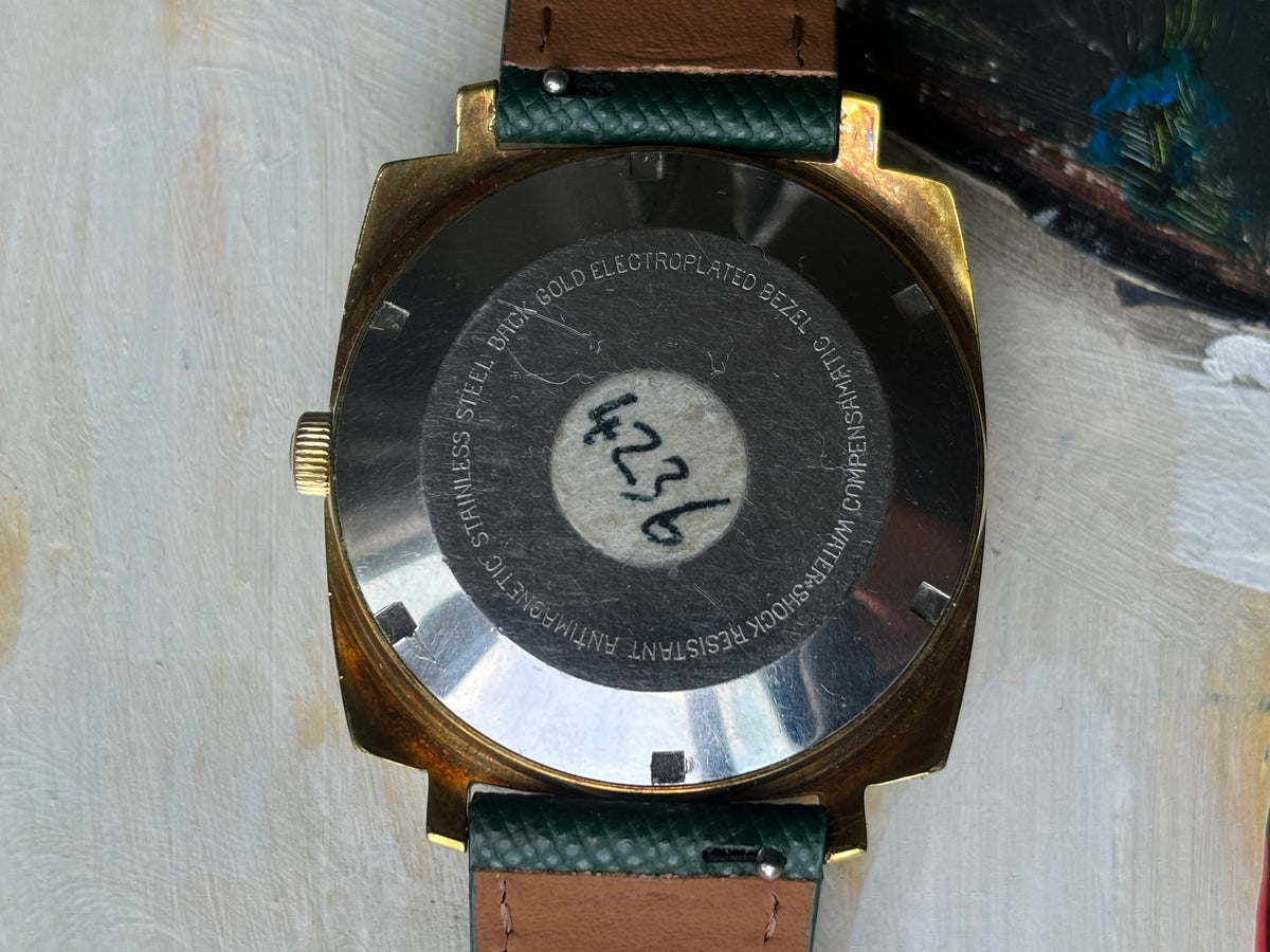 Close-up of the back of a wristwatch with a gold-tone case, brushed metal crystal cover, and a green-and-brown leather strap on a textured off-white surface.