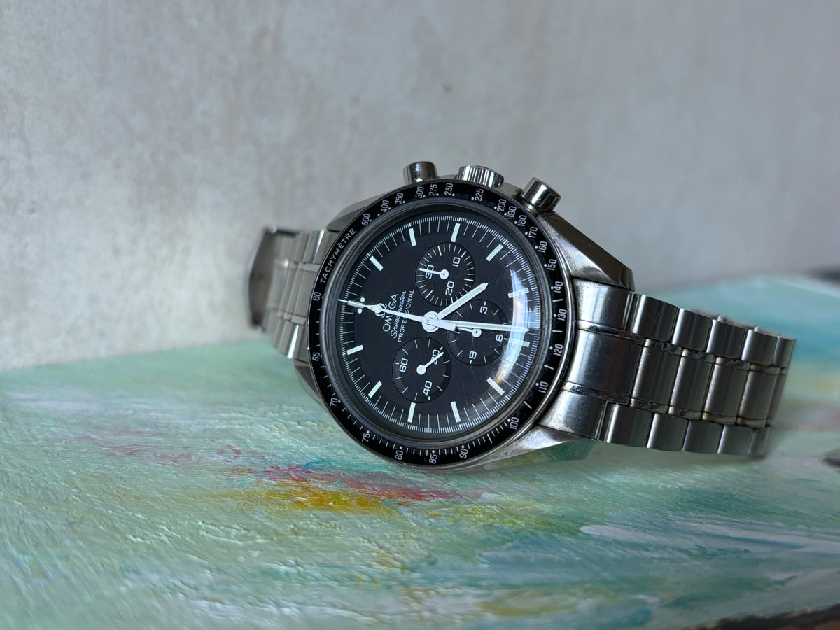 Omega Speedmaster Professional Moonwatch on a stainless-steel bracelet against a painted backdrop.